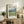 May include: Framed Van Gogh museum poster featuring swirling blue sky over green rolling hills and golden wheat fields, displayed on rustic wood sideboard with fern plant, ceramic vase, stacked books, and cream upholstered chair in neutral-toned living room setting.