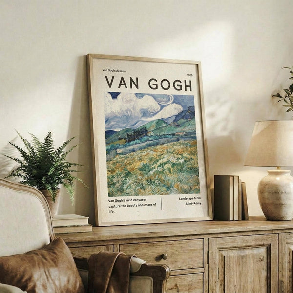 May include: Framed Van Gogh museum poster featuring swirling blue sky over green rolling hills and golden wheat fields, displayed on rustic wood sideboard with fern plant, ceramic vase, stacked books, and cream upholstered chair in neutral-toned living room setting.