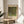 May include: Framed art print of Gustav Klimt's pointillist-style painting featuring a large tree with dense green and yellow foliage dotted with small white flowers, displayed on a round wooden side table next to a beige upholstered chair with white curtains and plant leaves visible in soft natural lighting.