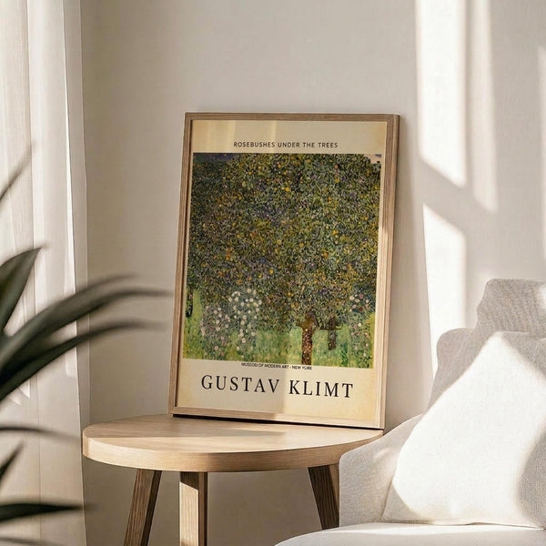 May include: Framed art print of Gustav Klimt's pointillist-style painting featuring a large tree with dense green and yellow foliage dotted with small white flowers, displayed on a round wooden side table next to a beige upholstered chair with white curtains and plant leaves visible in soft natural lighting.