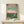 May include: Japanese woodblock print style poster in natural wood frame showing two cranes - one flying with outstretched wings against pale blue sky, another standing by water's edge. Scene features bare tree branches, distant pink sunset horizon, green marshland, and traditional Japanese text. Framed artwork leans against white wall on light wood surface next to small wrapped gift with twine and greenery sprig.
