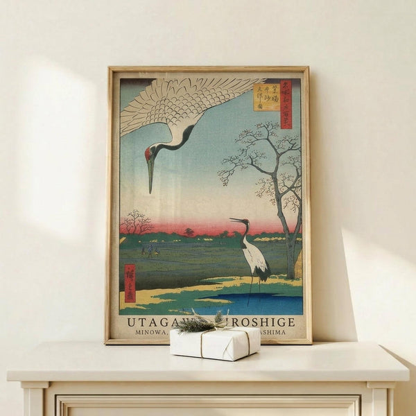 May include: Japanese woodblock print style poster in natural wood frame showing two cranes - one flying with outstretched wings against pale blue sky, another standing by water's edge. Scene features bare tree branches, distant pink sunset horizon, green marshland, and traditional Japanese text. Framed artwork leans against white wall on light wood surface next to small wrapped gift with twine and greenery sprig.