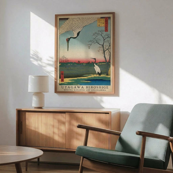 May include: Framed Japanese woodblock print showing two cranes in a landscape with water, trees, and mountains at sunset, displayed above a mid-century modern wooden sideboard with table lamp in a minimalist room with sage green upholstered chair and natural lighting casting shadows on white walls