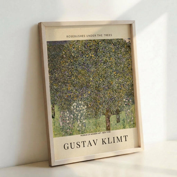 May include: Framed art print of Gustav Klimt's impressionistic painting featuring lush green foliage and rosebushes with delicate pink and white flowers beneath trees. The poster has cream-colored borders with black text reading "Rosebushes Under the Trees" at top and "Gustav Klimt" and "Museum of Modern Art - New York" at bottom. Light wood frame leaning against white wall.