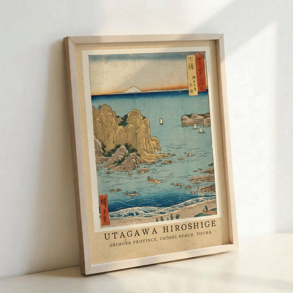 May include: Framed vintage Japanese woodblock print showing a coastal scene with rocky cliffs, blue ocean waters, small boats, and Mount Fuji in the distance. The artwork features traditional ukiyo-e style with muted earth tones and blue hues, displayed in a light wooden frame against a white wall. Japanese text cartouches visible in upper right and lower left corners of the print.