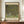 May include: Framed Gustav Klimt reproduction poster displayed on wooden credenza against cream paneled wall. The artwork shows flowering trees with dense green foliage dotted with colorful blooms in Klimt's distinctive pointillist style. Museum exhibition poster format with cream matting and text. Scene includes white ceramic vase and stacked books as styling props.