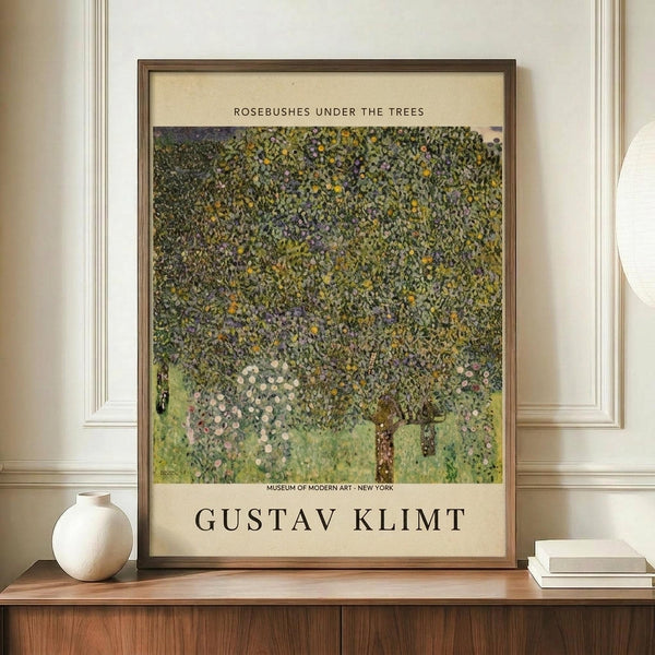 May include: Framed Gustav Klimt reproduction poster displayed on wooden credenza against cream paneled wall. The artwork shows flowering trees with dense green foliage dotted with colorful blooms in Klimt's distinctive pointillist style. Museum exhibition poster format with cream matting and text. Scene includes white ceramic vase and stacked books as styling props.