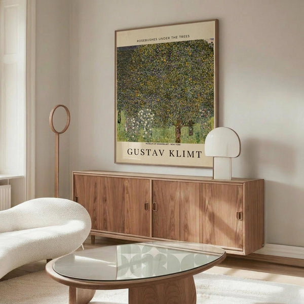 May include: Framed Gustav Klimt poster featuring "Rosebushes Under the Trees" artwork displayed above a wooden sideboard with sliding doors in a modern living room. The poster shows Klimt's distinctive pointillist style with dense green foliage and flowering trees. Room includes white curved chair, glass oval coffee table, modern white table lamp, and copper floor lamp against beige walls.