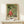 May include: Framed Japanese woodblock print featuring figures in traditional dress with animals including a tiger, set against green geometric background. Light wood frame sits on pale wooden surface beside small white wrapped gift. Minimalist neutral wall setting with soft natural lighting.