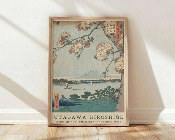 May include: Framed Japanese woodblock print reproduction leaning against white wall on light wood floor. Artwork shows cherry blossoms framing view of Mount Fuji across water with boats, featuring traditional ukiyo-e style in muted blues, greens, and pink tones. Natural wood frame with white matting, soft natural lighting creates diagonal shadows on wall.