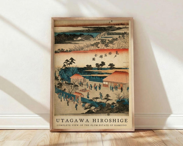 May include: Framed Japanese woodblock print showing traditional landscape scene with buildings, people in traditional dress, water features, and trees in muted colors including teal, coral, and beige. Print displays in light wood frame leaning against white wall on light wooden floor with natural lighting casting soft shadows.