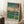 May include: Framed Japanese woodblock print reproduction featuring cherry blossom trees overlooking Mount Fuji, green fields, and blue river with small figures. Traditional ukiyo-e art style with vibrant blues, greens, and coral pink blossoms. Light wood frame leaning against white wall corner beside coffee cup on wooden block and cream textured rug.