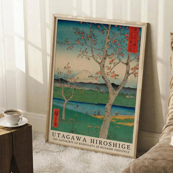 May include: Framed Japanese woodblock print reproduction featuring cherry blossom trees overlooking Mount Fuji, green fields, and blue river with small figures. Traditional ukiyo-e art style with vibrant blues, greens, and coral pink blossoms. Light wood frame leaning against white wall corner beside coffee cup on wooden block and cream textured rug.
