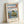 May include: A framed Japanese woodblock print reproduction in a light wooden frame, leaning against a white wall. The artwork depicts dramatic rocky sea stacks rising from blue ocean waters, with a red torii gate perched on top of the largest rock formation. Mount Fuji appears as a distant silhouette on the horizon under a pale sky with orange sunset tones. Traditional Japanese text cartouches appear in the upper right and lower left corners. The print shows classic ukiyo-e style with bold colo