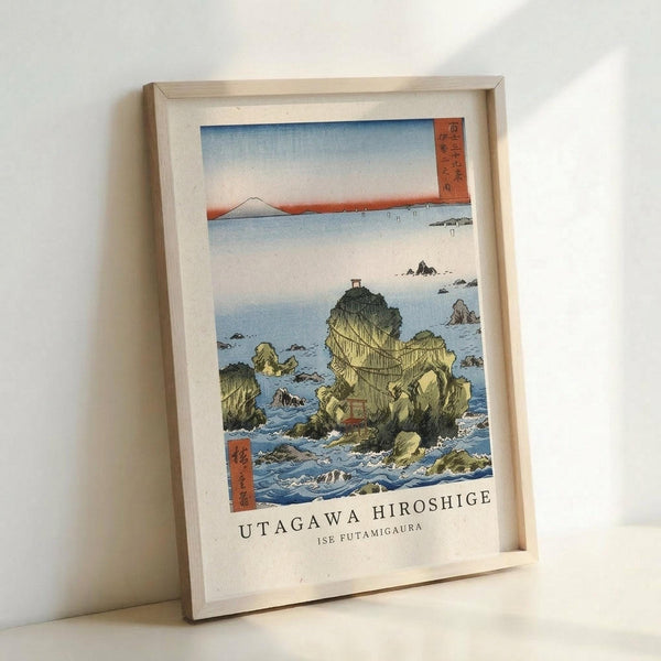 May include: A framed Japanese woodblock print reproduction in a light wooden frame, leaning against a white wall. The artwork depicts dramatic rocky sea stacks rising from blue ocean waters, with a red torii gate perched on top of the largest rock formation. Mount Fuji appears as a distant silhouette on the horizon under a pale sky with orange sunset tones. Traditional Japanese text cartouches appear in the upper right and lower left corners. The print shows classic ukiyo-e style with bold colo