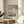 May include: Japanese woodblock print poster in light wooden frame displayed on round wooden side table next to beige upholstered chair. Artwork shows cherry blossoms framing view of Mount Fuji across water with boats, featuring soft blues, greens, and pink tones. Natural sunlight streams across white wall creating geometric shadows. Green plant leaves visible in corner.
