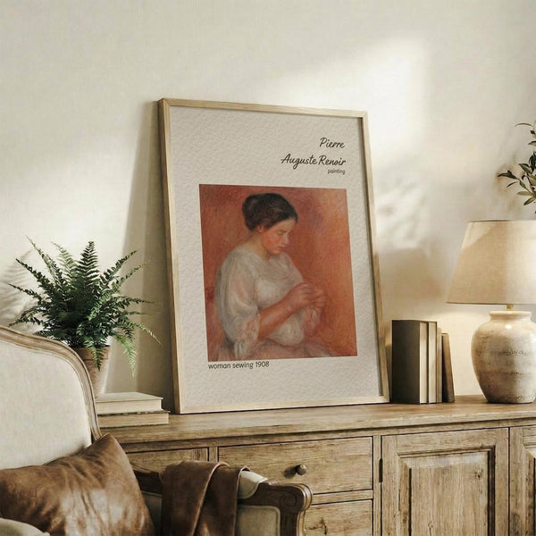 May include: Framed reproduction of Renoir's Woman Sewing 1908 displayed on rustic wooden credenza in neutral living room. Print shows woman in white dress against warm orange background. Scene includes fern plant, stacked books, ceramic vase, table lamp, and beige upholstered chair with brown throw pillow. Natural lighting creates soft shadows on cream walls.