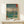 May include: Japanese woodblock print poster in wooden frame showing cherry blossom tree beside blue river with Mount Fuji in background, displayed on light wood console table next to small wrapped gift box against white wall