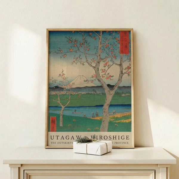 May include: Japanese woodblock print poster in wooden frame showing cherry blossom tree beside blue river with Mount Fuji in background, displayed on light wood console table next to small wrapped gift box against white wall