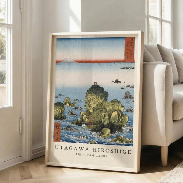 May include: Framed Japanese woodblock print showing rocky coastal cliffs jutting from blue ocean waters with Mount Fuji visible on distant horizon under pale sky. Print features traditional ukiyo-e art style with red Japanese text cartouches, displayed in light wood frame leaning against white wall next to beige upholstered sofa in bright modern living room with large windows.