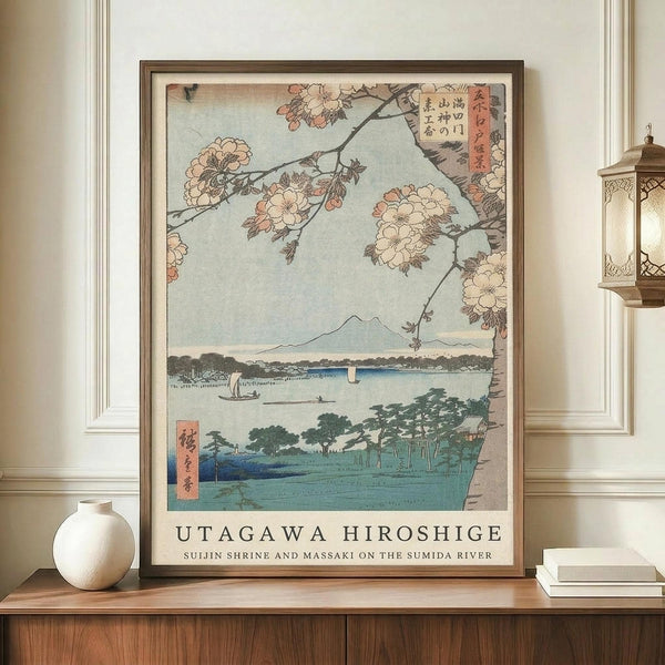 May include: Framed Japanese woodblock print displaying cherry blossoms overhanging a serene river landscape with Mount Fuji in the distance, small boats on blue water, and traditional architecture. The artwork features soft pink and white blossoms against a pale blue sky, with green hills and trees below. Displayed on a warm wood sideboard alongside a cream ceramic vase and stacked books, with an ornate brass lantern visible on the right side of the elegant room setting.