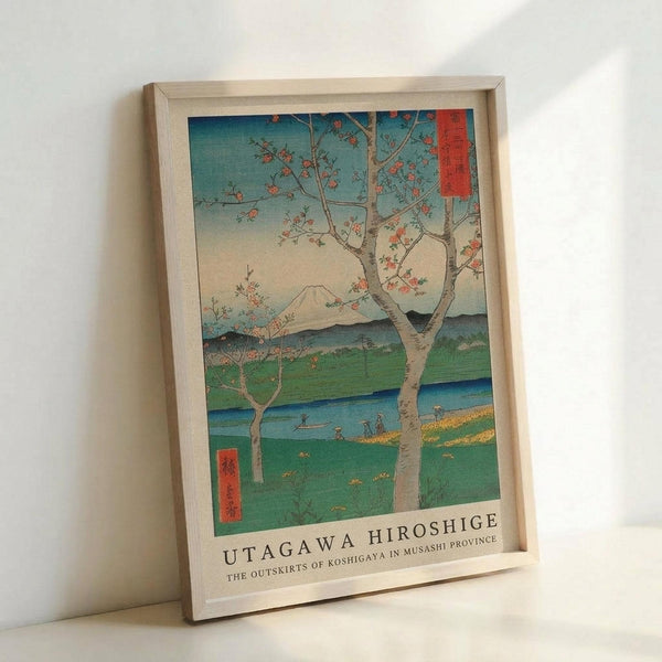 A framed reproduction of a Japanese woodblock print showing a flowering tree in the foreground with pink blossoms, overlooking a green landscape with a winding blue river and distant mountains. The artwork features the characteristic ukiyo-e style with vibrant blues, greens, and coral tones, plus red text cartouches with Japanese characters. The print is displayed in a natural wood frame against a white wall, with "UTAGAWA HIROSHIGE" and subtitle text printed below the image.