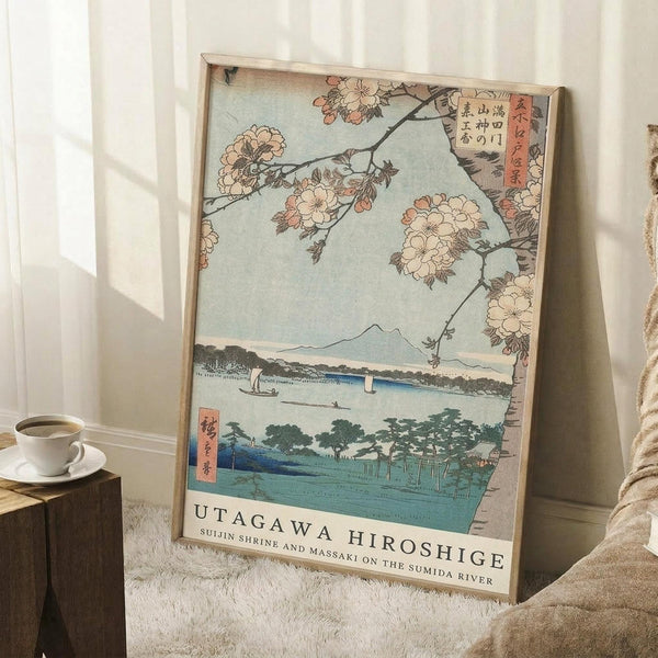 May include: Japanese woodblock print reproduction in light wooden frame leaning against white wall corner. Features cherry blossoms framing Mount Fuji view across blue water with traditional boats and green landscape. Soft natural lighting creates shadows on white textured rug. Coffee cup on wooden block visible nearby for scale. Beige throw blanket partially shown.