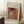 May include: Framed art print leaning against white wall on textured cream rug, showing Renoir's soft-toned painting of woman in white blouse sewing, with warm orange background. Light wood frame with cream matting. Coffee cup on wooden block nearby, cozy home setting with natural window light creating shadows.