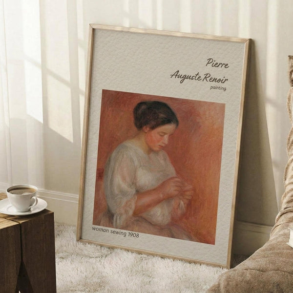 May include: Framed art print leaning against white wall on textured cream rug, showing Renoir's soft-toned painting of woman in white blouse sewing, with warm orange background. Light wood frame with cream matting. Coffee cup on wooden block nearby, cozy home setting with natural window light creating shadows.
