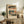 May include: Framed Japanese woodblock print artwork displayed on rustic wooden sideboard in neutral-toned living room. Print features traditional landscape scenes with blue water, orange buildings, small figures, and Japanese text, shown in light wooden frame. Scene includes fern plant, ceramic vase, books, and cream upholstered furniture.