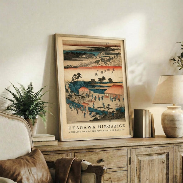 May include: Framed Japanese woodblock print artwork displayed on rustic wooden sideboard in neutral-toned living room. Print features traditional landscape scenes with blue water, orange buildings, small figures, and Japanese text, shown in light wooden frame. Scene includes fern plant, ceramic vase, books, and cream upholstered furniture.