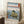 May include: Framed Japanese woodblock print poster leaning against white wall corner, featuring coastal rocky formations with traditional shrine torii gate, blue ocean waves, and Mount Fuji in distant background. Print shows classic ukiyo-e art style with green and yellow rock formations, blue water, and text at bottom. Small wooden side table with white coffee cup visible on left, beige throw blanket draped nearby on white textured rug.