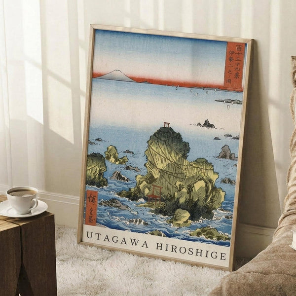 May include: Framed Japanese woodblock print poster leaning against white wall corner, featuring coastal rocky formations with traditional shrine torii gate, blue ocean waves, and Mount Fuji in distant background. Print shows classic ukiyo-e art style with green and yellow rock formations, blue water, and text at bottom. Small wooden side table with white coffee cup visible on left, beige throw blanket draped nearby on white textured rug.
