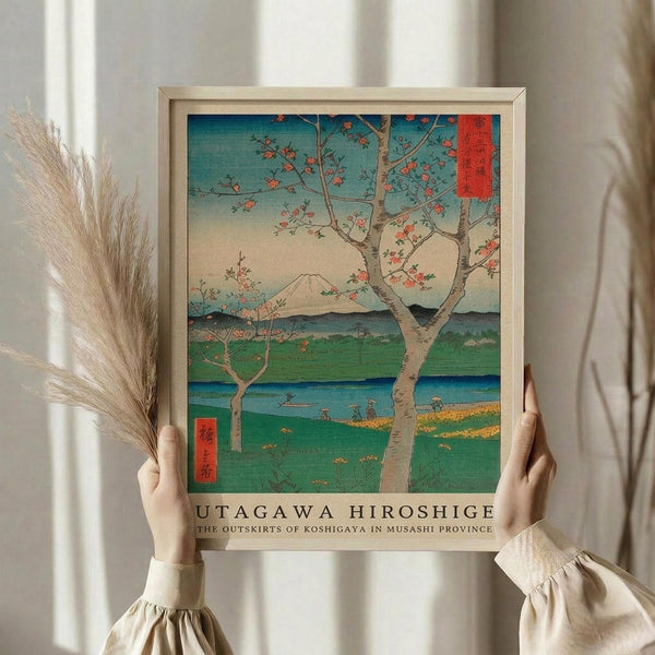 May include: Person in cream clothing holding framed Japanese woodblock print featuring cherry blossom tree with pink flowers, Mount Fuji in background, green fields and blue water, traditional ukiyo-e art style with turquoise sky. Light wood frame, dried pampas grass visible beside figure against soft neutral curtained background.