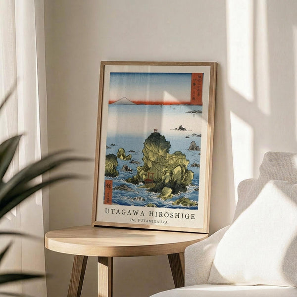 May include: Framed Japanese woodblock print showing rocky coastal formations jutting from blue ocean waters with Mount Fuji visible in distance under gradient sky. Print displays traditional ukiyo-e art style with green and yellow rocky outcrops, small boats, and red cartouche with Japanese text. Natural wood frame sits on round wooden side table next to beige upholstered chair and plant leaves in bright, sunlit room with white walls.