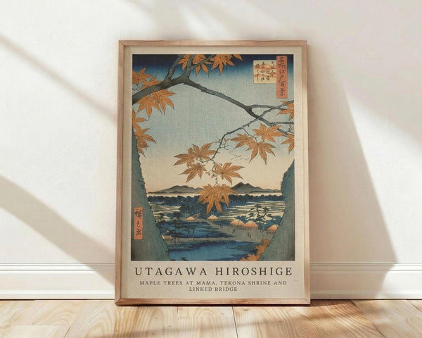 May include: Framed Japanese woodblock print reproduction showing autumn maple branches with orange leaves overhanging a blue river landscape with mountains in the distance. The artwork features traditional ukiyo-e style with Japanese text cartouches. Natural wood frame leaning against white wall on light wooden floor with soft natural lighting creating diagonal shadows.