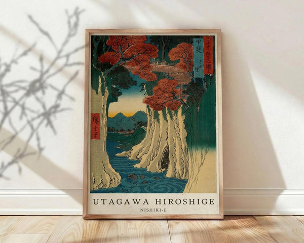May include: Framed Japanese woodblock print showing dramatic waterfall cascading down rocky cliffs with red autumn maple trees above, traditional pagoda structure visible in upper portion, blue river flowing below, mountains in distance under gradient sky, wooden frame leaning against white wall on light wooden floor with plant shadow visible.