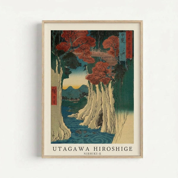 Framed Japanese woodblock print showing a dramatic waterfall cascading down white cliffs into blue water, with vibrant orange-red maple trees above and mountains in the distance. The artwork features traditional ukiyo-e style with Japanese text characters in the upper right corner. Displayed in a light wood frame against a white wall, the print has rich teal, orange, and cream colors with detailed natural scenery.