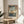 May include: Japanese woodblock print artwork displayed in light wooden frame on round wooden side table next to beige upholstered chair. Print shows autumn maple branches with orange leaves overlooking blue water and mountains, featuring traditional ukiyo-e style with Japanese text. Soft natural lighting creates shadows on neutral wall, with plant leaves visible in corner.