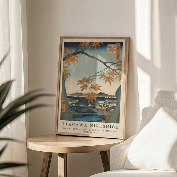 May include: Japanese woodblock print artwork displayed in light wooden frame on round wooden side table next to beige upholstered chair. Print shows autumn maple branches with orange leaves overlooking blue water and mountains, featuring traditional ukiyo-e style with Japanese text. Soft natural lighting creates shadows on neutral wall, with plant leaves visible in corner.