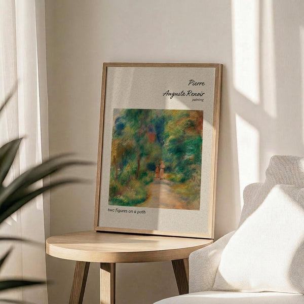 May include: Framed art poster featuring an impressionist forest path painting with lush green foliage and two small figures walking along a winding trail. The print displays "Pierre Auguste Renoir painting" and "two figures on a path" text on cream paper within a thin gold frame, placed on a round wooden side table next to a beige upholstered chair with natural lighting casting shadows across the neutral-toned room.