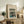 May include: Framed Japanese woodblock print reproduction showing fishing boats with nets on blue water at dusk, displayed on rustic wooden sideboard alongside fern plant, ceramic vases, and books in warm-toned living room setting with upholstered chair visible.