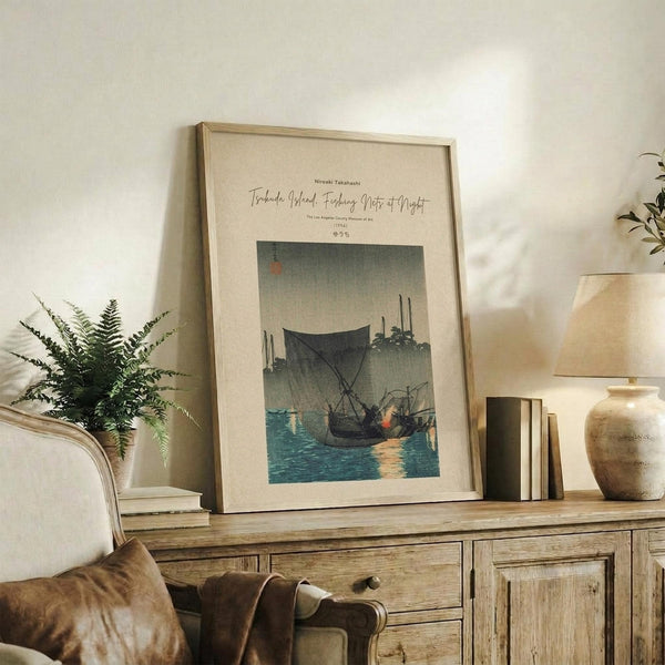 May include: Framed Japanese woodblock print reproduction showing fishing boats with nets on blue water at dusk, displayed on rustic wooden sideboard alongside fern plant, ceramic vases, and books in warm-toned living room setting with upholstered chair visible.