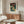 May include: Japanese woodblock print poster featuring autumn trees with red foliage, white cliffs, and blue water, displayed in thin wooden frame above mid-century walnut credenza. Modern living room setting with white upholstered chair, glass-top oval coffee table, and minimal lighting on neutral walls.