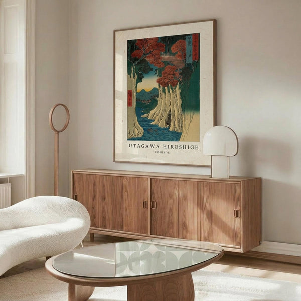 May include: Japanese woodblock print poster featuring autumn trees with red foliage, white cliffs, and blue water, displayed in thin wooden frame above mid-century walnut credenza. Modern living room setting with white upholstered chair, glass-top oval coffee table, and minimal lighting on neutral walls.
