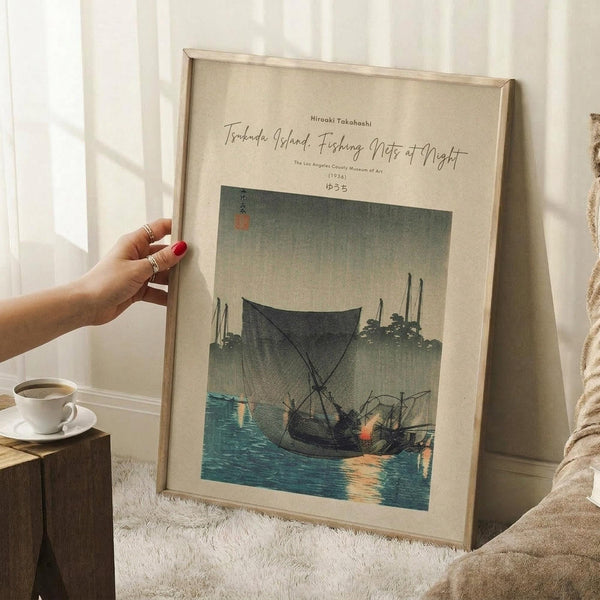 May include: A framed Japanese woodblock print showing fishing boats with nets on blue water at dusk, held by a hand with red nail polish. The artwork features muted greens and blues with traditional calligraphy. Scene includes a coffee cup on wooden surface and white textured rug in soft natural lighting.