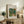 May include: Framed art print of Renoir's impressionist painting showing two small figures walking on a woodland path surrounded by lush green foliage in blues, greens, and earth tones. The poster sits on a rustic wooden credenza alongside books, a fern plant, and ceramic vase in a neutral-toned living room setting with natural lighting.