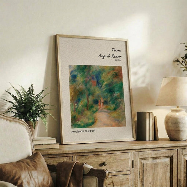 May include: Framed art print of Renoir's impressionist painting showing two small figures walking on a woodland path surrounded by lush green foliage in blues, greens, and earth tones. The poster sits on a rustic wooden credenza alongside books, a fern plant, and ceramic vase in a neutral-toned living room setting with natural lighting.