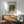 May include: Framed Renoir reproduction print showing impressionist landscape painting with two figures on garden path surrounded by lush green foliage. Print displayed above mid-century modern wood credenza next to ceramic table lamp, with mint green upholstered armchair in foreground. Warm natural lighting creates geometric shadows on white wall.