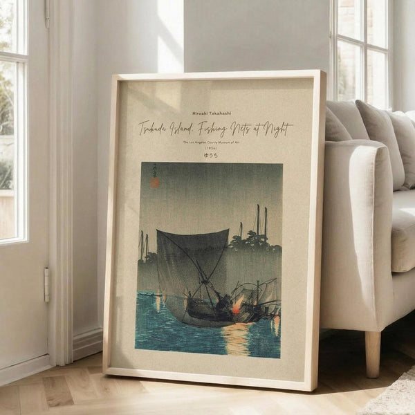 May include: Framed poster print leaning against white wall next to beige upholstered chair in bright living room with large windows. The artwork depicts traditional Japanese woodblock print style scene with fishing boats on teal water at dusk, featuring dark silhouettes against muted green-gray sky. Print has cream colored background with black text above the image. Natural wood frame, approximately 2-3 feet tall, casting soft shadows on light wooden floor.